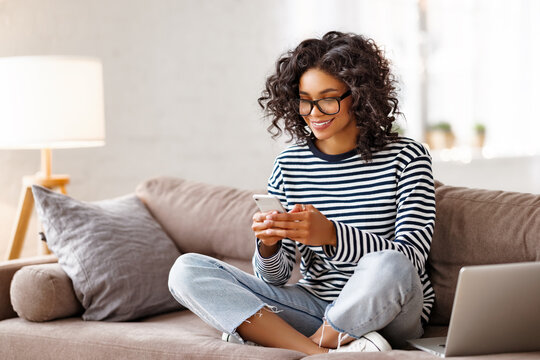 Cheerful ethnic female using smartphone on couch