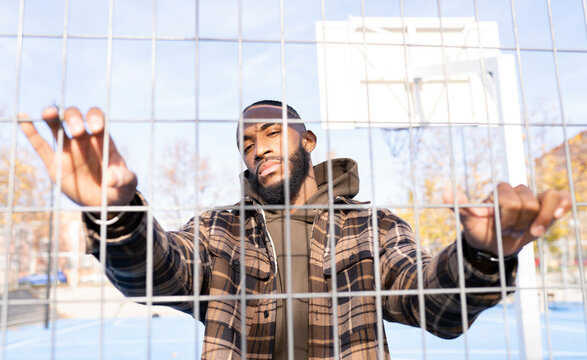 Young man with cool attitude standing behind fence in sports court on sunny day