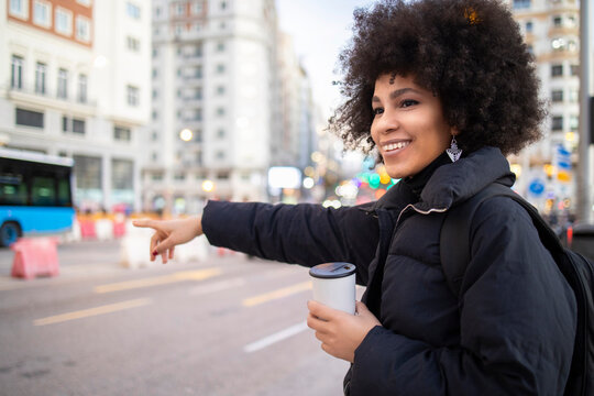 Afro young woman holding disposable coffee cup while hailing taxi on city street