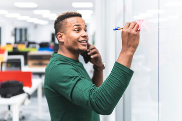 Smiling businessman talking on mobile phone while writing on adhesive note in office corridor