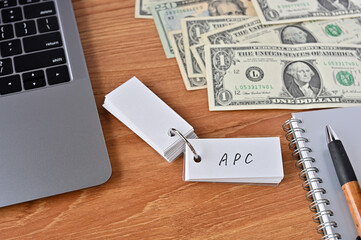 On the desk there are bills, a laptop, and a word book with the word APC written on it. It was an abbreviation for the financial term average propensity to consume.