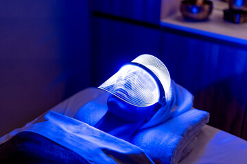 Young woman wearing illuminated blue mask during rejuvenating treatment in dark at beauty spa