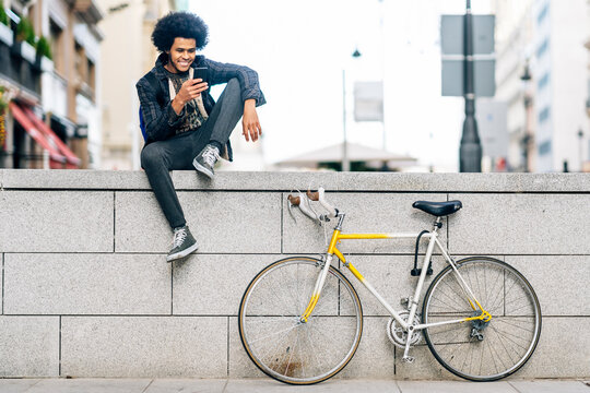 Young man with curly hair using smart phone while sitting on retaining wall