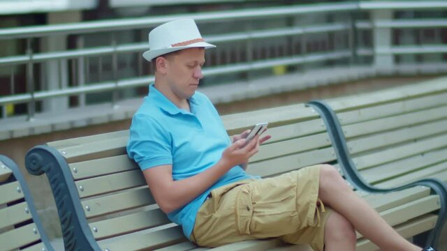 Young man in white hat sits on a city bench with a mobile phone in his hands, camera tracking