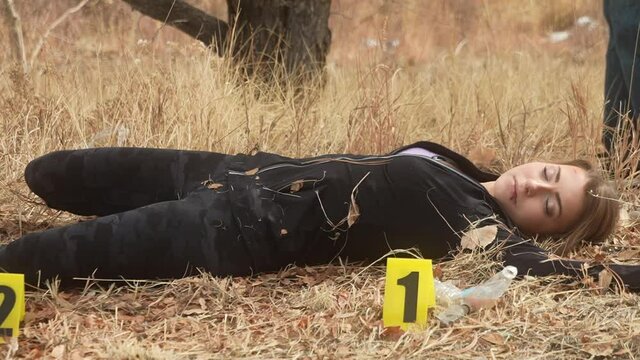 Deceased female crime victim surrounded by evidence markers examined by detective for evidence behind police tape on grass field