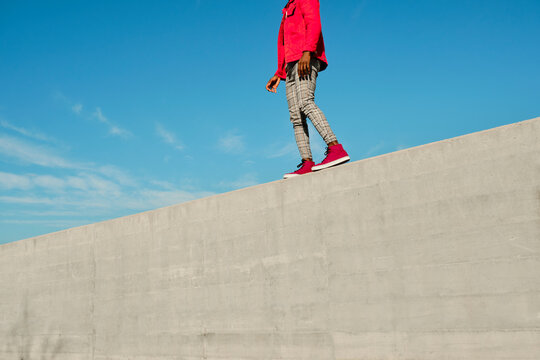 Young man in red jacket and checked pants walking on concrete wall