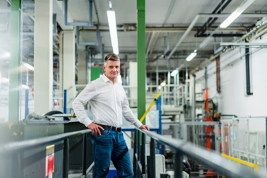 Smiling mature businessman with hand on hip standing by railing in factory