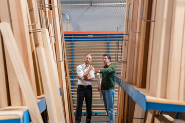 Two carpenters talking in production hall with wooden planks in foreground