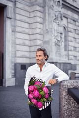 Fototapeta premium A handsome stylish man in a white shirt and black trousers and a bouquet of pink peonies in his hands waiting for his beloved girl.