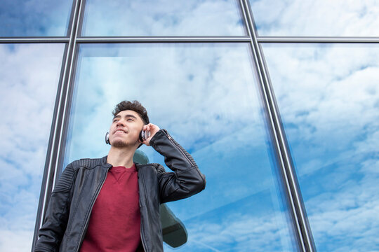 Smiling young man in leather jacket listening music through headphones against glass building