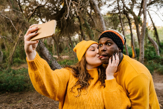 Young woman kissing man while taking selfie through mobile phone while standing at forest