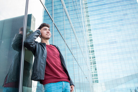 Smiling young man listening music through headphones while leaning on glass wall