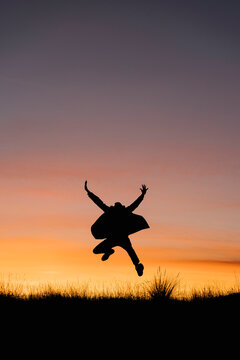Carefree man wearing jacket jumping with arms outstretched against sky during sunset