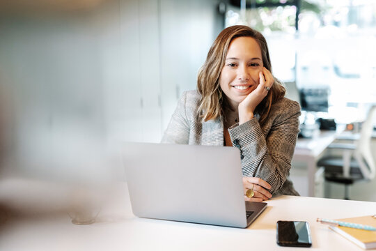 Smiling businesswoman with laptop on desk sitting in office - Powered by Adobe