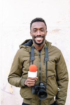 Smiling male reporter with microphone and camera standing against white wall