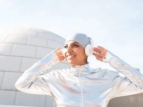 Smiling woman wearing protective suit listening music while standing against igloo