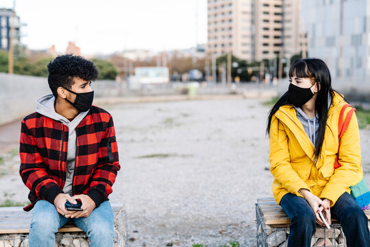 Couple Wearing Protective Face Mask Sitting At Social Distance On Bench During COVID-19