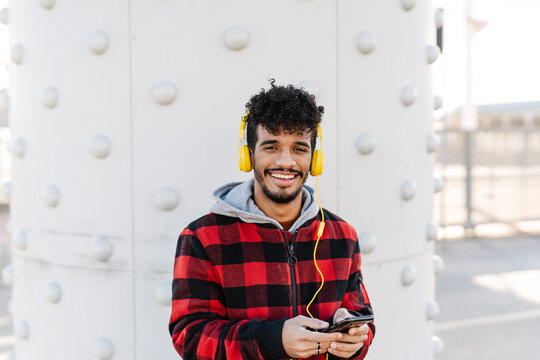 Man wearing hooded shirt listening music through headphones while standing with mobile phone against wall