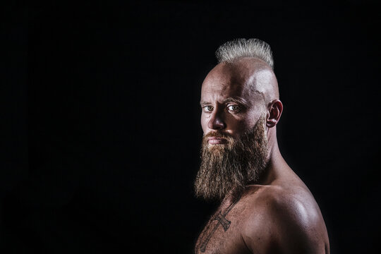 Shirtless male viking with beard and mohawk hairstyle against black background