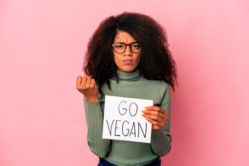 Young african american curly woman holding a go vegan placard showing fist to camera, aggressive facial expression.