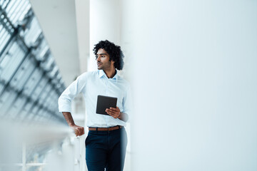 Thoughtful male professional holding digital tablet while standing by railing looking away in corridor at office