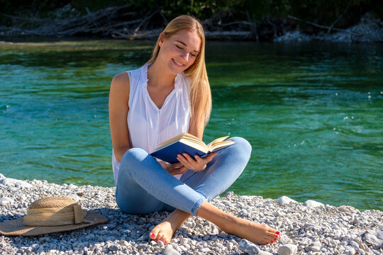 Smiling blond woman sitting cross-legged while reading book by river