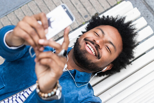 Close-up of cheerful man with afro hair listening music and using mobile phone while lying on bench