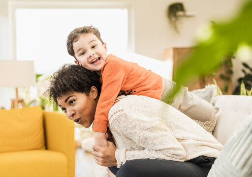 Playful Woman Piggybacking Cheerful Boy While Sitting On Sofa At Home