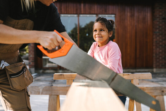 Smiling daughter looking at mother cutting wooden plank with hand saw against house
