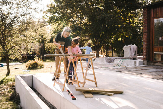 Mother cutting wood with hand saw by daughter outside house