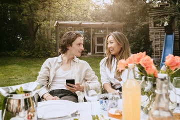 Smiling man with smart phone talking to female friend while sitting at table in backyard