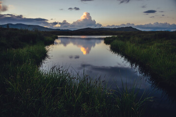Fototapeta premium Yellow big cloud in form of explosion above mountain lake near green grass on foreground. Awesome giant cloud reflected in lake water in sunrise. Huge cloud of illuminating color in dawn gradient sky.