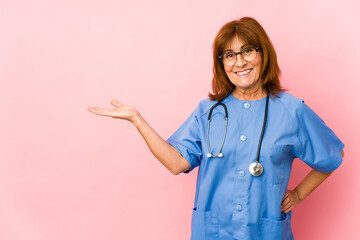 Middle age caucasian nurse woman isolated showing a copy space on a palm and holding another hand on waist.