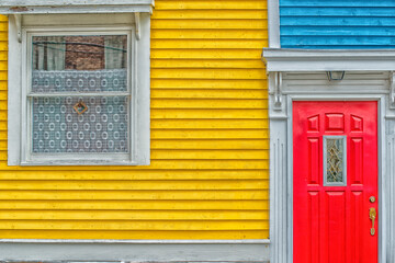 Fototapeta premium A bright red door of a building with blue and yellow wooden clapboard walls. The house has a red decorative metal mailbox. There's a green shrub near the red door with white trim. 