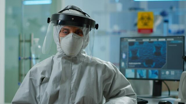 Tired microbiologist in coverall sitting in laboratory looking at camera. Team of scientits examining virus evolution using high tech and chemistry tools for scientific research, vaccine development.