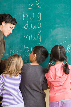 Children (6-7) writing on blackboard with teacher looking at them