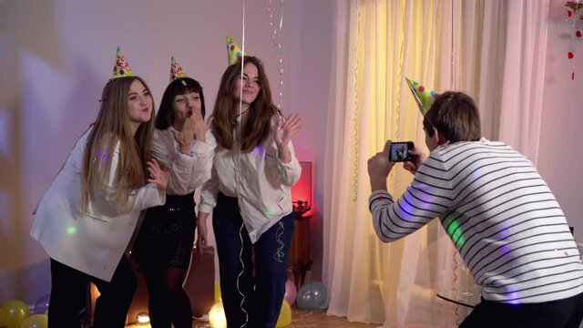 Millennial Caucasian man taking photos of cheerful young women on birthday party. Group of cheerful positive friends having fun partying at home indoors. Lifestyle and fun.