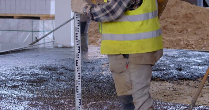 On a construction site, concrete is poured from a tractor bucket onto a cement screed, one worker align it, another checks the surface level and makes a mark on the concrete 
