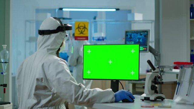 Scientist man wearing protection suit typing on computer with green mockup in modern equipped lab. Team of microbiologists doing vaccine research writing on device with chroma key, isolated display.