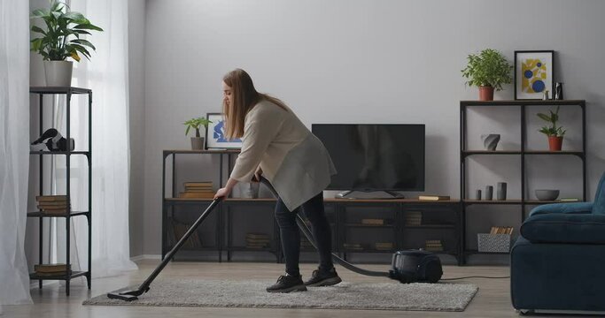 young woman is using vacuum cleaner in living room, cleaning home at weekend, housewife is doing housework