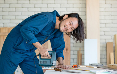 Good looking Asian carpenter wearing coverall dress working for DIY jobs in carpenter room with several kinds of woods and types of equipment