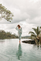 Happy young woman in long  dress walking on the edge of swimming pool and enjoying summer vacation in daytime