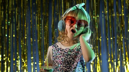 Futuristic hipster teen girl listening to music in headphones, dancing in slow motion at disco party cyberpunk club. Child kid in neon lights with pineapple fruit juice standing by illuminated wall