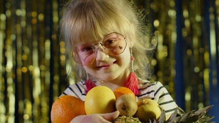 Close-up shot of girl in stylish sunglasses posing looking at camera with bunch of fruits in hands on shiny background in night club. Child kid sensual look clubber in neon yellow lights