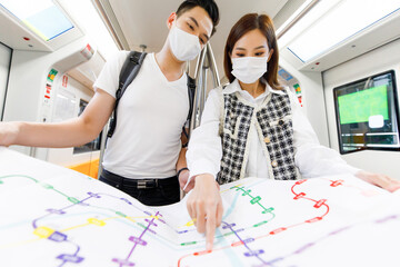 Front view portrait of young adult Asian couple tourists with a medical mask standing and holding the paper metro map together in the Skytrain with a blurred map foreground and Skytrain background