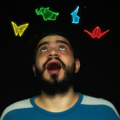 Fondo negro con un hombre joven de buso a rayas azul con blanco, barba y cabello negro mira sobre él una mariposa, elefante, conejo y una grulla hechos en origami mágico flotando con destellos de luz © Diego