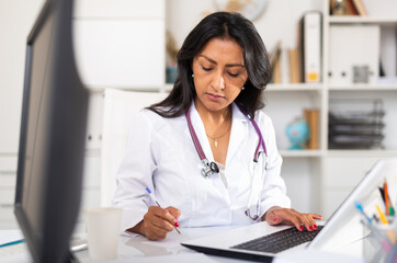 Latin american woman experienced physician filling up medical forms on laptop while sitting at table in office