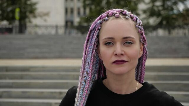 Unusual young woman with colored pigtails and bright make-up mysteriously looks into the camera against the backdrop of the cityscape. High quality FullHD footage