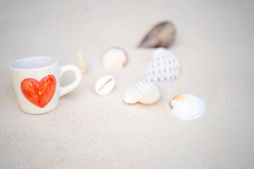 cup coffee with red heart printed and shells on sandy background.
