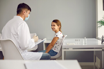 Man doctor pediatrician in medical uniform and mask talking with little smiling girl patient and...